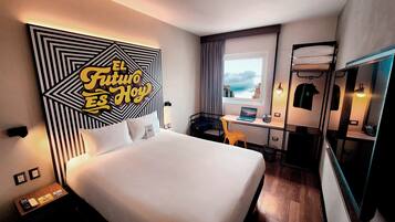 Habitación de hotel con una cama grande, un escritorio con una computadora portátil, un espejo y vistas a la ciudad.