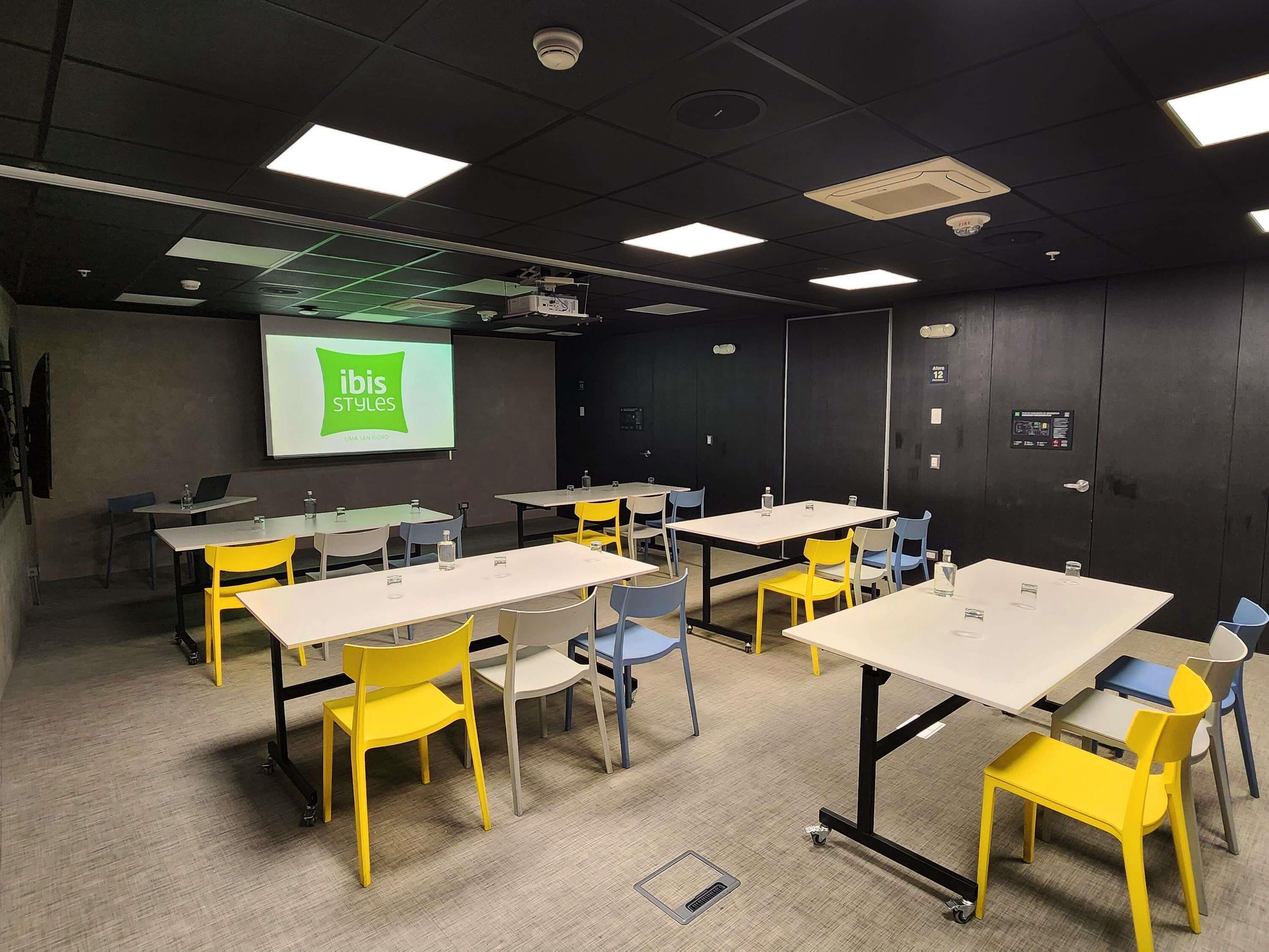 A conference room with tables and chairs, a projector screen displaying 'ibis Styles,' and a fire alarm on the wall.