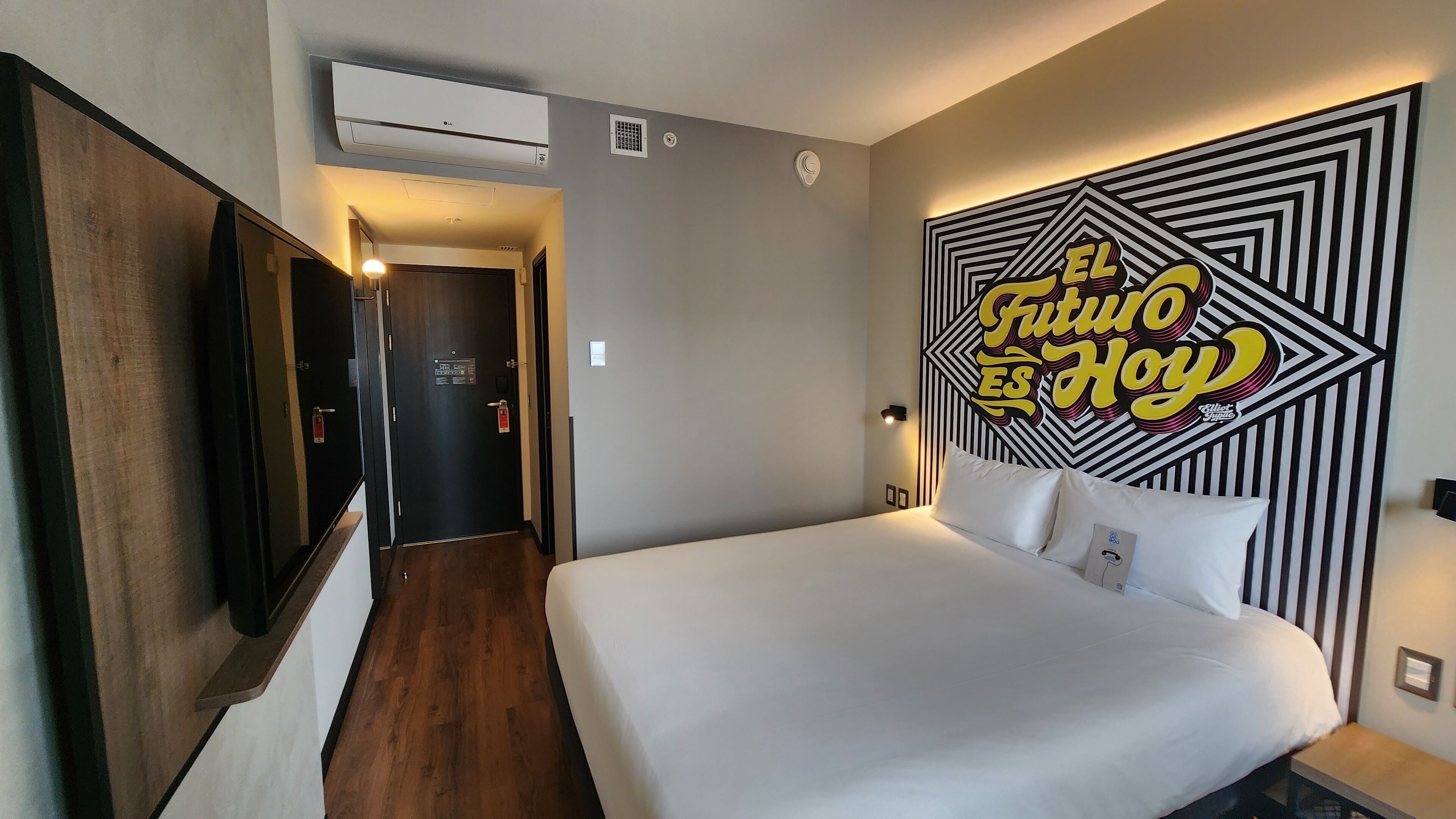 Una habitación de hotel con una cama, aire acondicionado montado en la pared, una puerta con un letrero y un adorno geométrico en la pared.