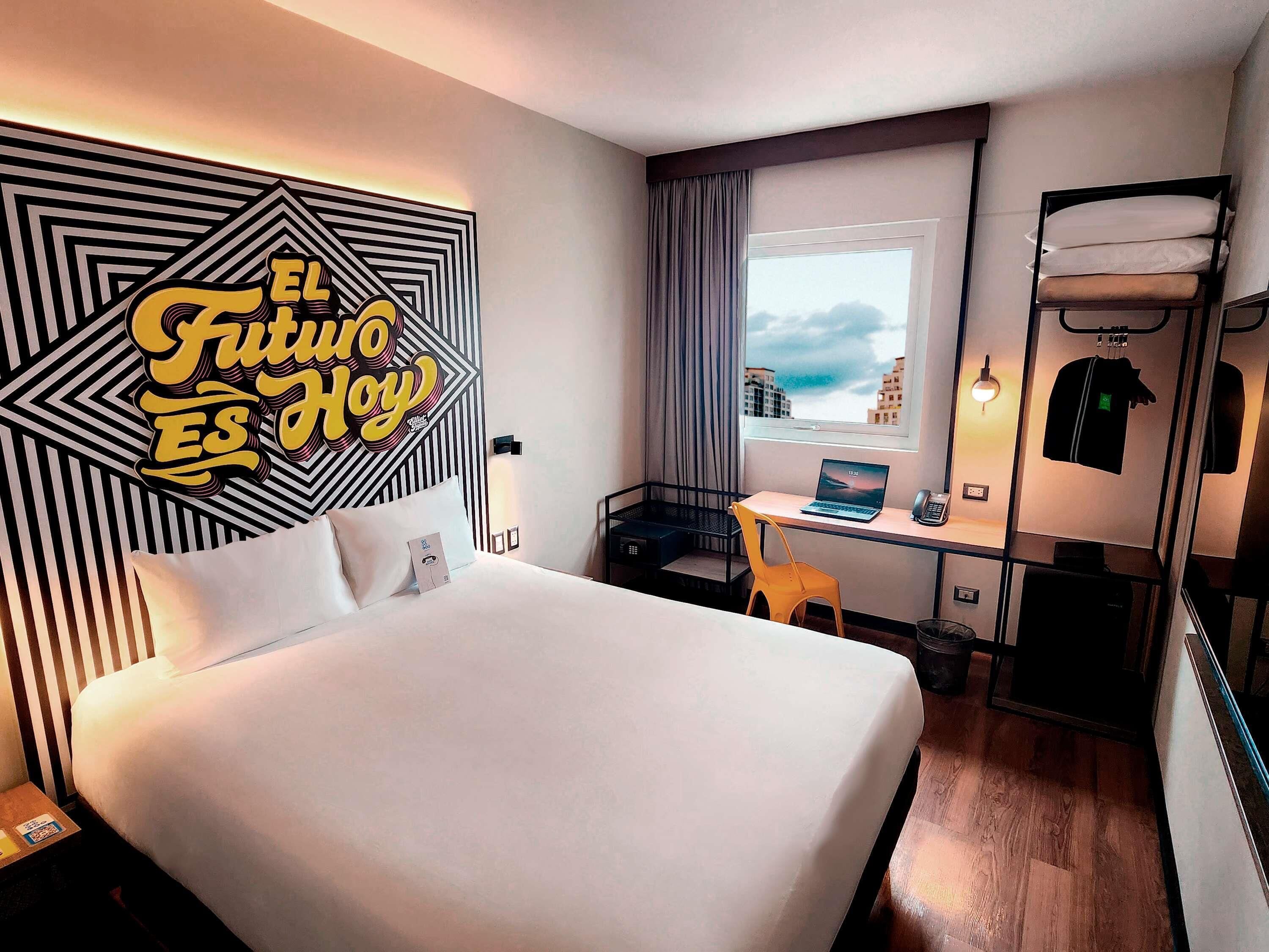 A hotel room with a bed, a desk with a laptop, a chair, a window with a view, and a wall with a geometric pattern.