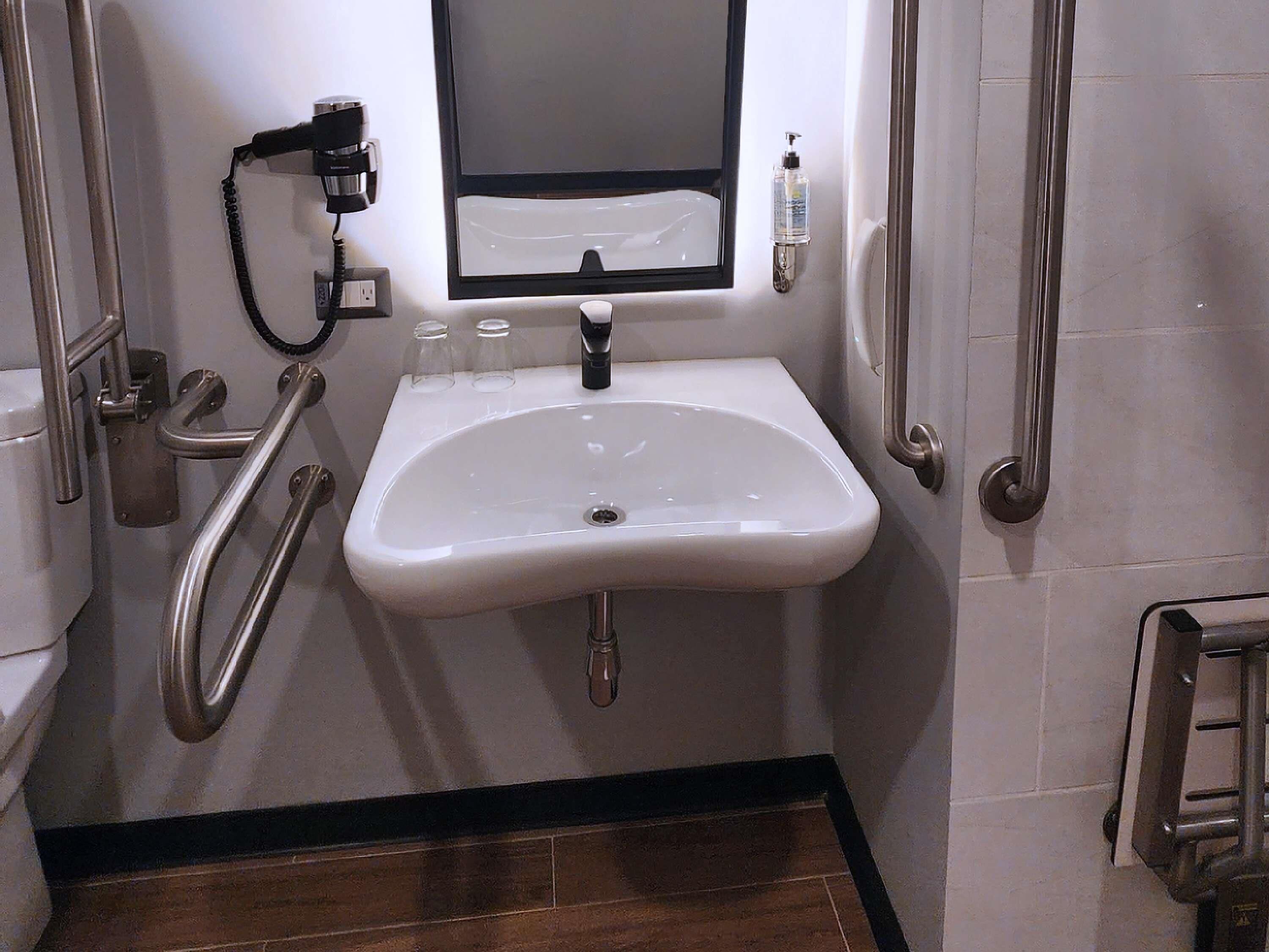 A bathroom with a white sink, a mirror, a hand dryer, and a hand sanitizer dispenser.