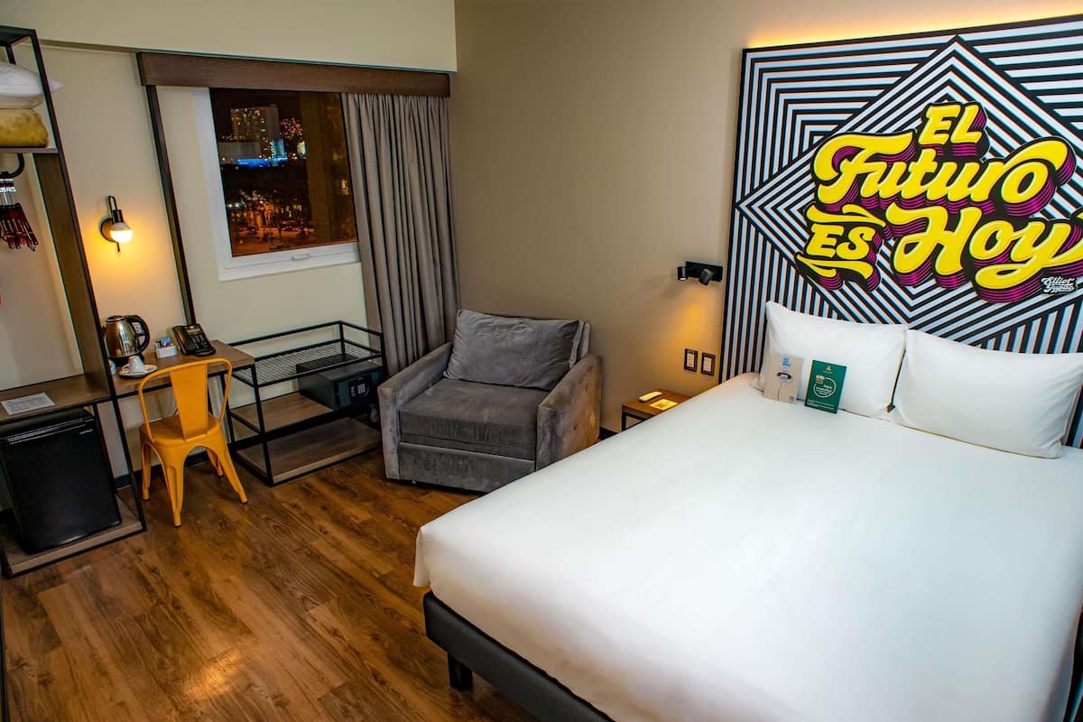 Habitación de hotel con cama, escritorio, silla, mesita pequeña, mesita de noche y ventana con cortinas.
