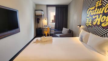 Habitación de hotel con una cama grande, un escritorio con silla, una mesita, un televisor y una pared con diseño geométrico.