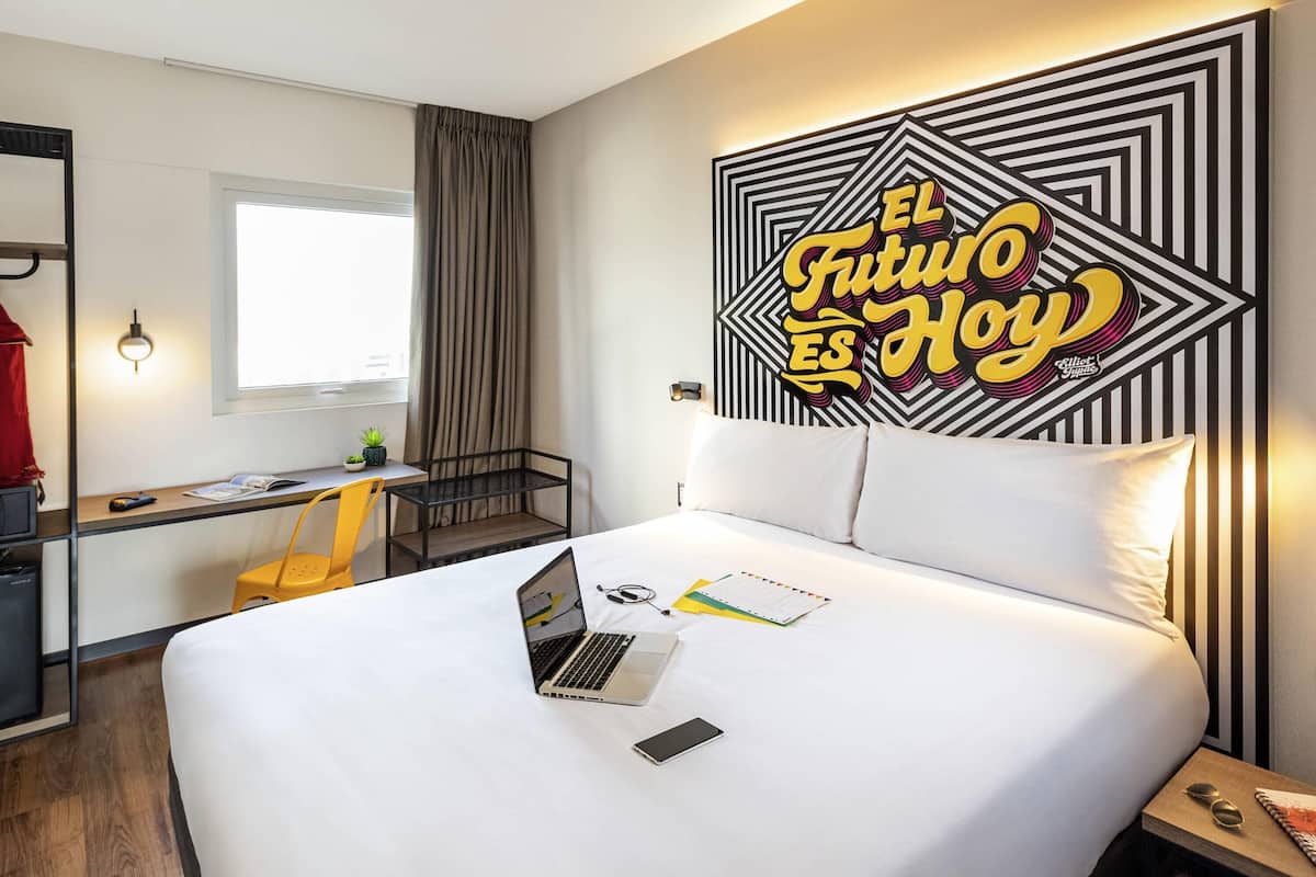 Una habitación de hotel con una cama, un escritorio con una computadora portátil, una silla y una pared con un diseño geométrico.