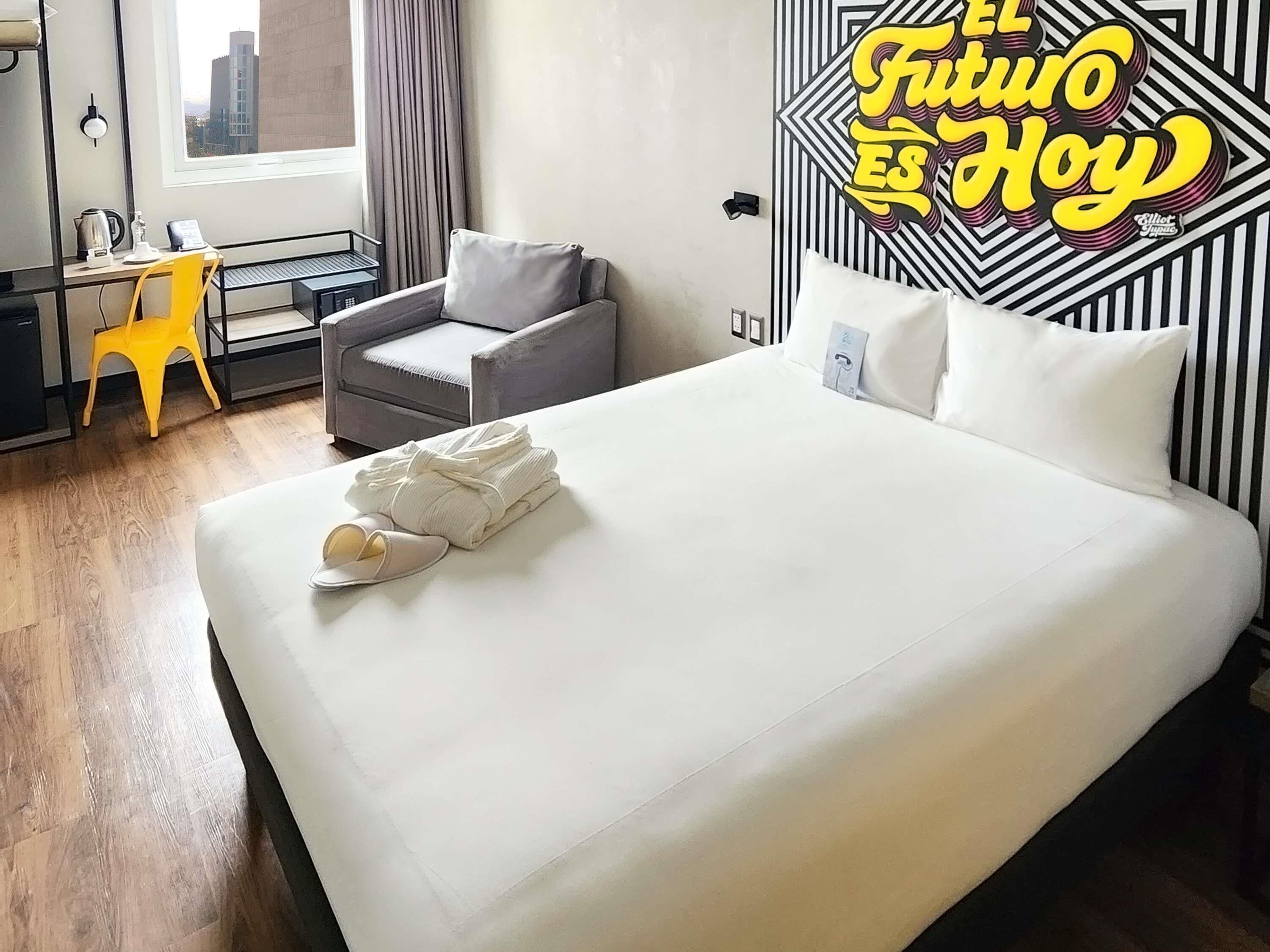 Una habitació d'hotel amb un llit, una cadira, un escriptori i una finestra.