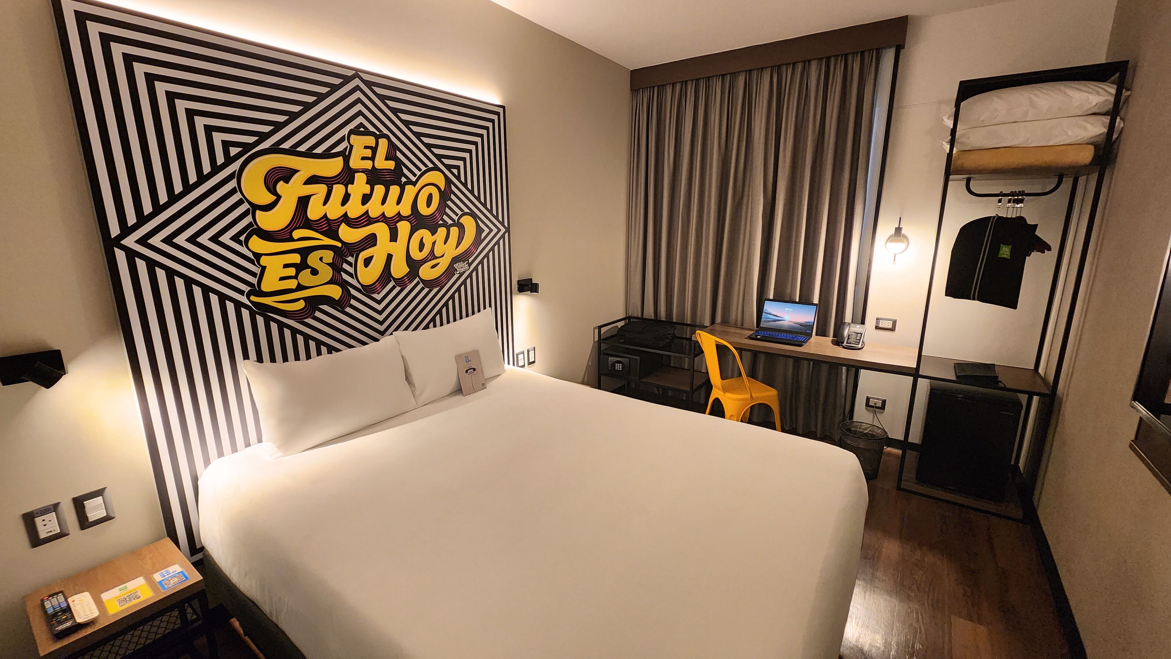 A hotel room with a large bed, a desk with a laptop, a chair, a small table with a phone, and a wall with a geometric design.
