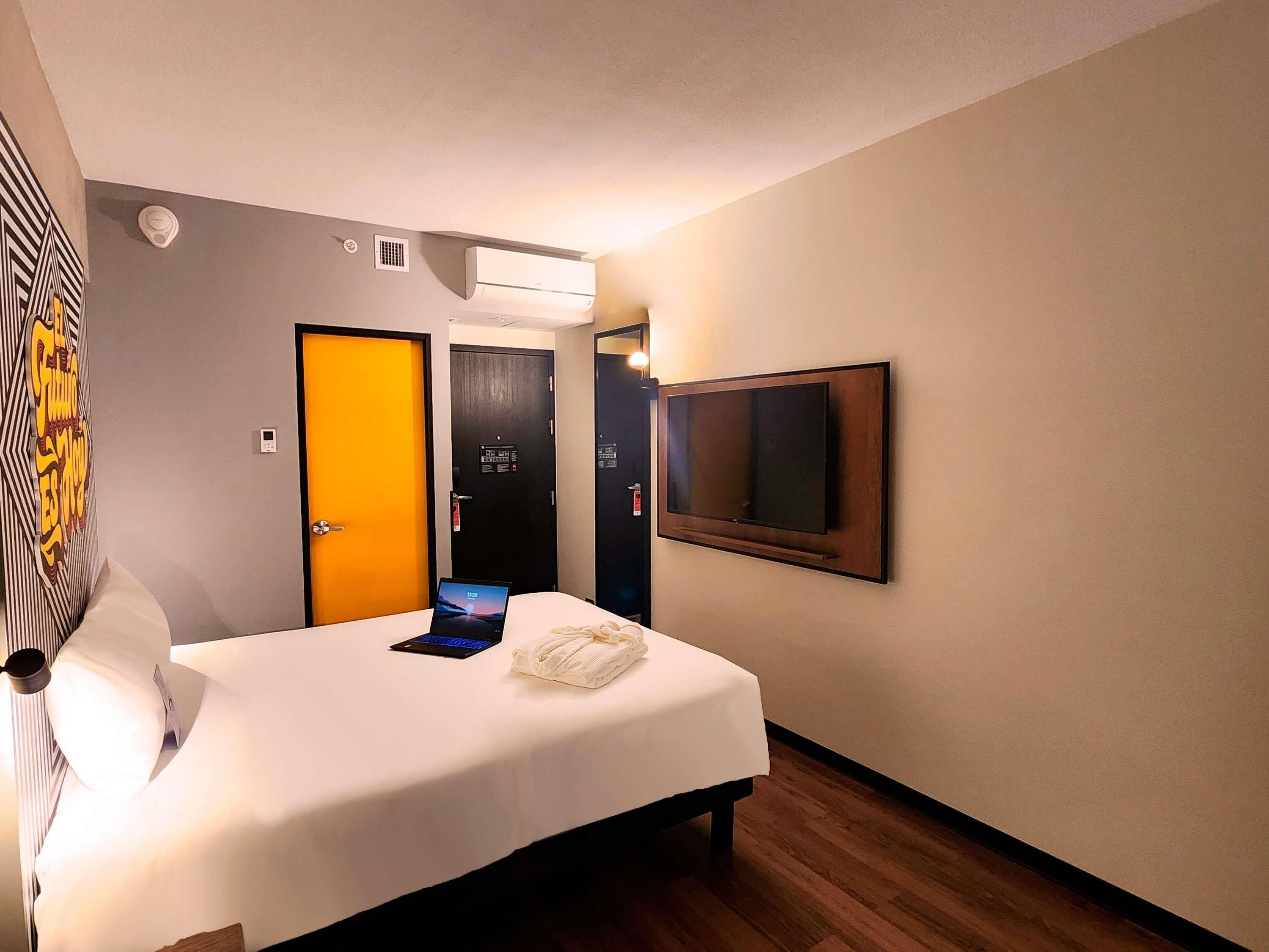 A hotel room with a bed, a flat-screen TV mounted on the wall, an air conditioning unit, and a door with a digital display.