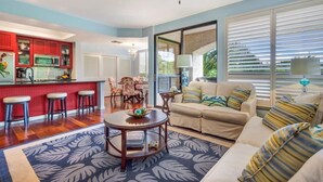A spacious living area with a sofa, a coffee table, and a kitchen area with red cabinets and a bar. - Comfortable and cozy condo (Waikoloa Village)