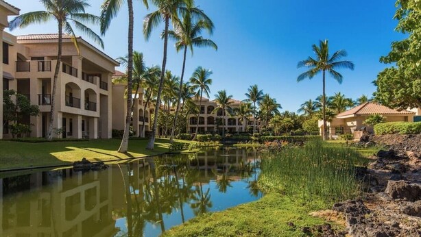 A resort with multiple-story buildings, a central water feature, and palm trees. - Comfortable and cozy condo (Waikoloa Village)