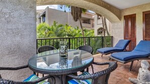 A balcony with wicker furniture, a glass table, and a view of greenery and buildings. - Comfortable and cozy condo (Waikoloa Village)