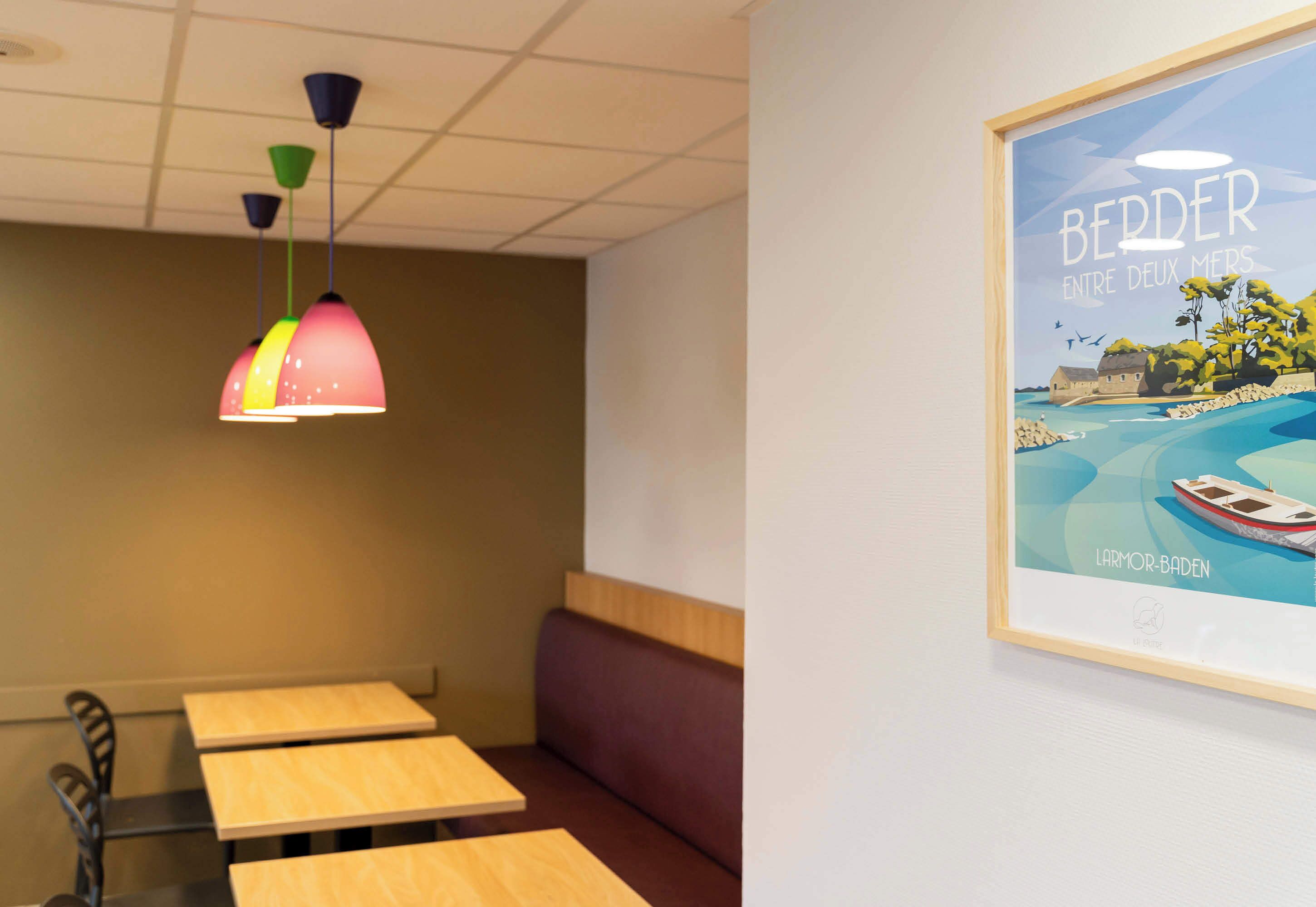 A room with wooden tables and benches, colorful pendant lights, and a framed picture of a coastal scene.