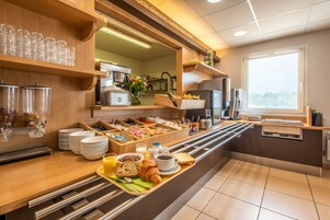 A breakfast buffet with various food items and beverages, a coffee machine, and a window with blinds.