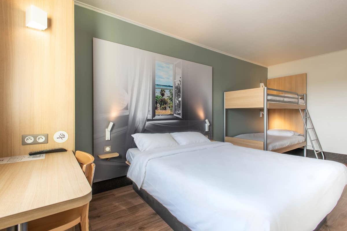A hotel room with a bunk bed, a desk with a chair, a large window with a view, and a wall-mounted light fixture.