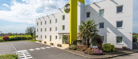 A modern hotel with a prominent yellow accent, a parking area, and a well-maintained garden.