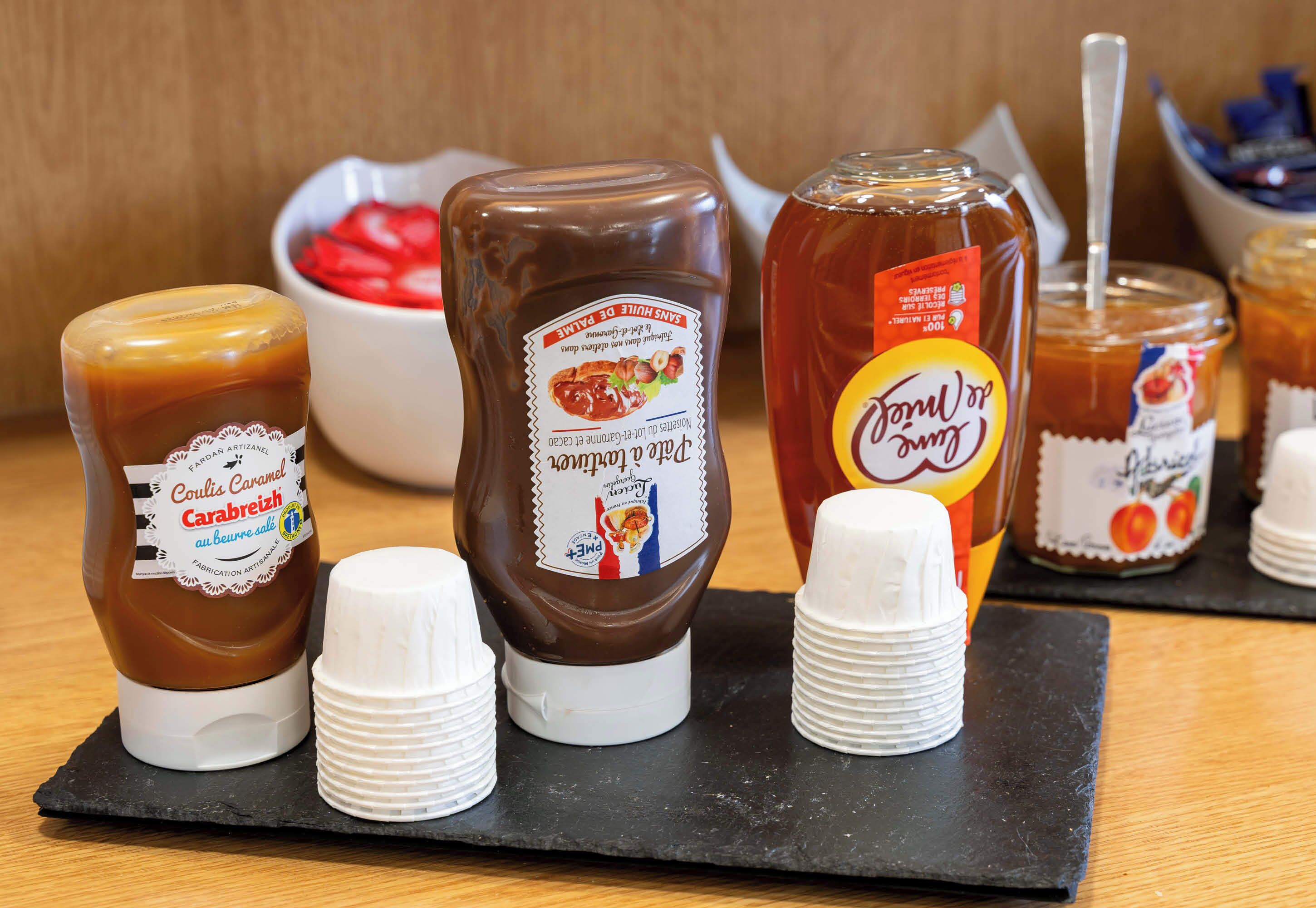 Variedad de condimentos, incluyendo salsa de caramelo, mostaza y mostaza con miel, dispuestos sobre una tabla de pizarra negra con vasos de papel blancos.
