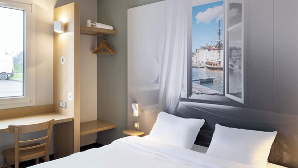 A hotel room with a bed, a desk, a chair, a wardrobe, and a large window with a cityscape view. - B&B HOTEL Vannes Ouest Golfe du Morbihan (Vannes)