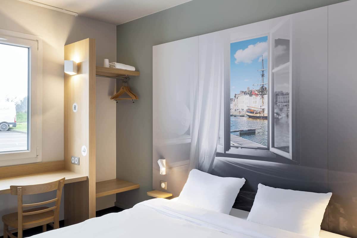 A hotel room with a bed, a desk, a chair, a wardrobe, and a large window with a cityscape view.
