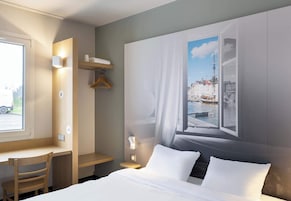A hotel room with a bed, a desk, a chair, a wardrobe, and a large window with a cityscape view.