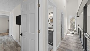 Un pasillo con una puerta blanca, un televisor de pantalla plana montado en la pared, y una cocina visible a través de la puerta.