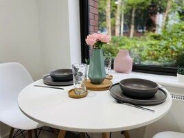 A round dining table with a white tablecloth, black plates, and cutlery. There is a teal vase with pink flowers, a clear glass, and a pink vase on the table.