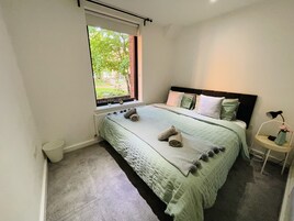 Un dormitorio con un ventanal grande, una cama con cubrecama verde y una mesita de noche con lámpara.