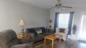Un salon avec un canapé gris, une table basse en bois, un fauteuil gris, un ventilateur de plafond et une fenêtre avec des stores.