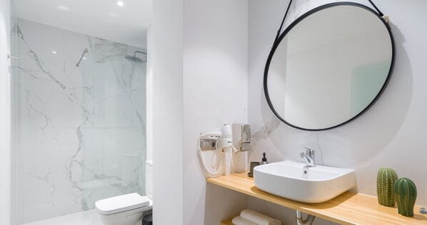 Un baño moderno con ducha de mármol, lavamanos blanco, espejo redondo y un cactus en la encimera.