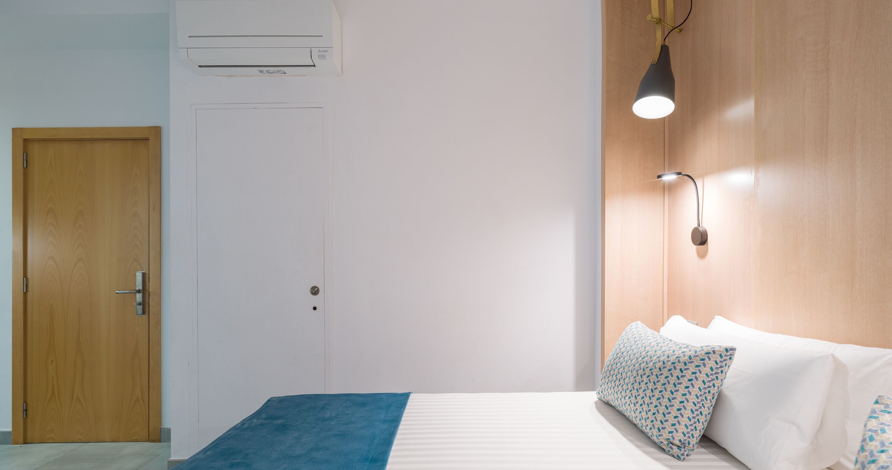 Habitación de hotel con puerta de madera, cama con ropa de cama blanca y almohadas estampadas, una manta azul y una lámpara fijada a la pared.