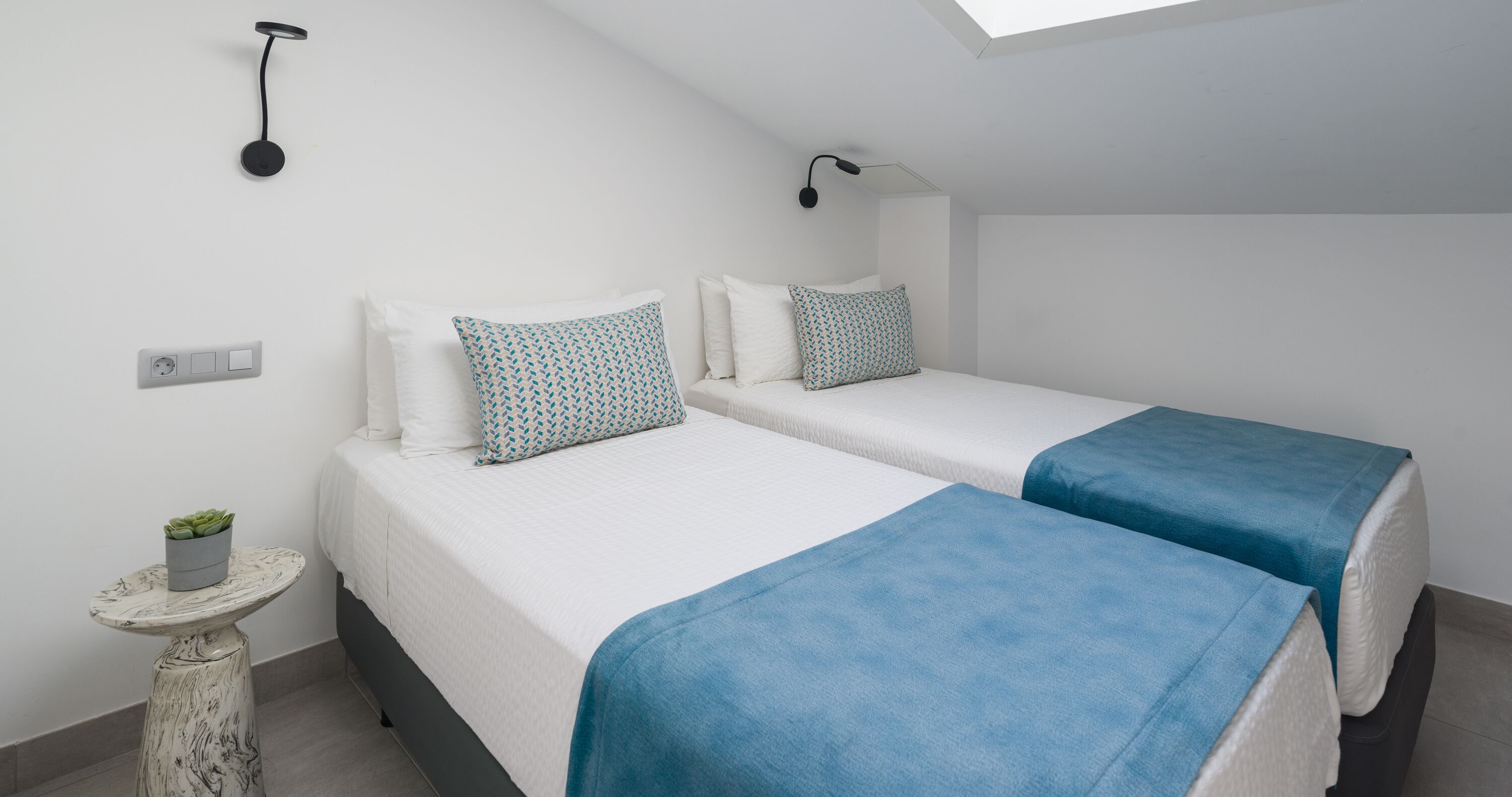 A bedroom with two beds, a small table with a plant, a wall socket, and a ceiling light fixture.