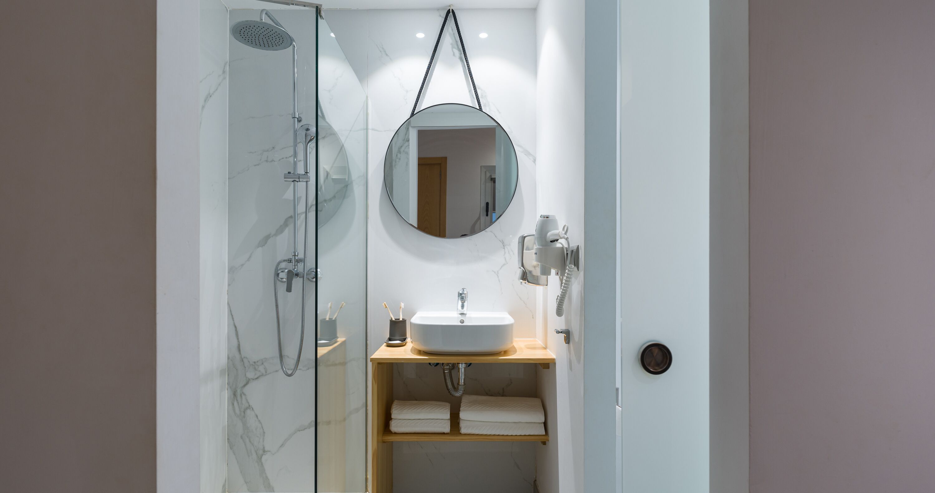 A bathroom with a glass-enclosed shower, a round mirror, a white sink, and a wooden shelf with towels.