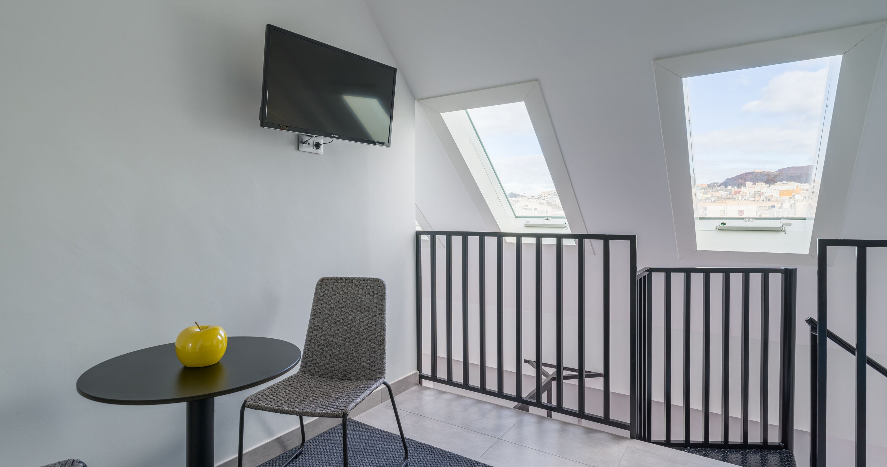 A modern room with a flat-screen TV mounted on the wall, a small round table with a yellow apple, a grey patterned chair, and a view through large windows to the cityscape.