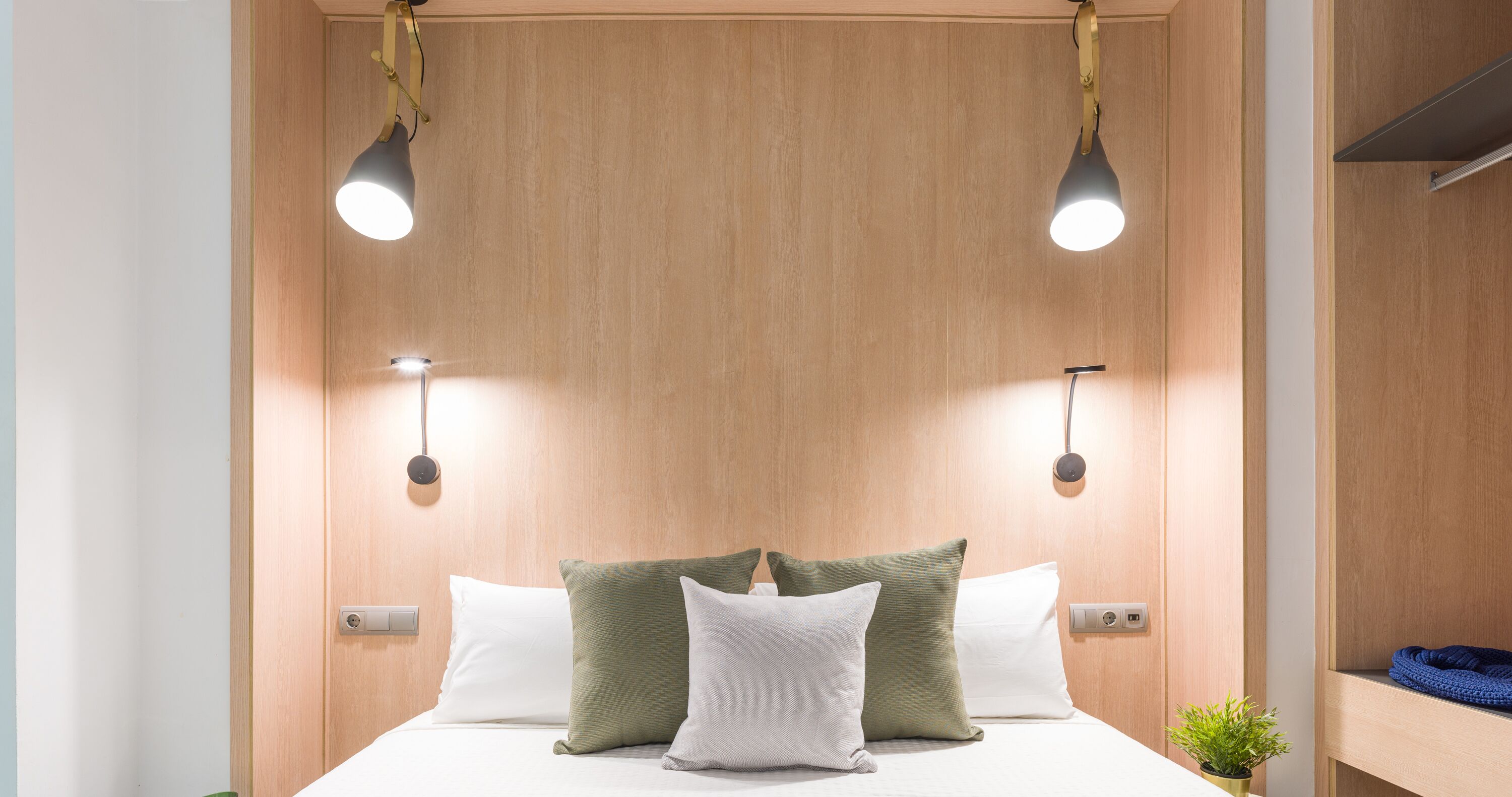 A neatly made bed with white and green pillows, two wall-mounted lights, and a small plant on the side.