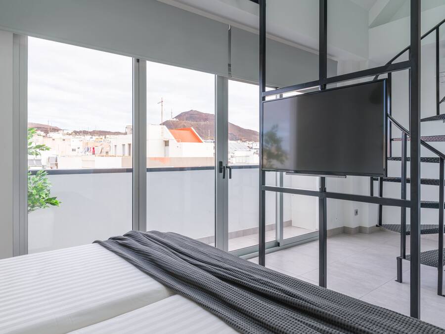 A modern bedroom with a large bed, a TV, and a balcony with a view of buildings and a hill.