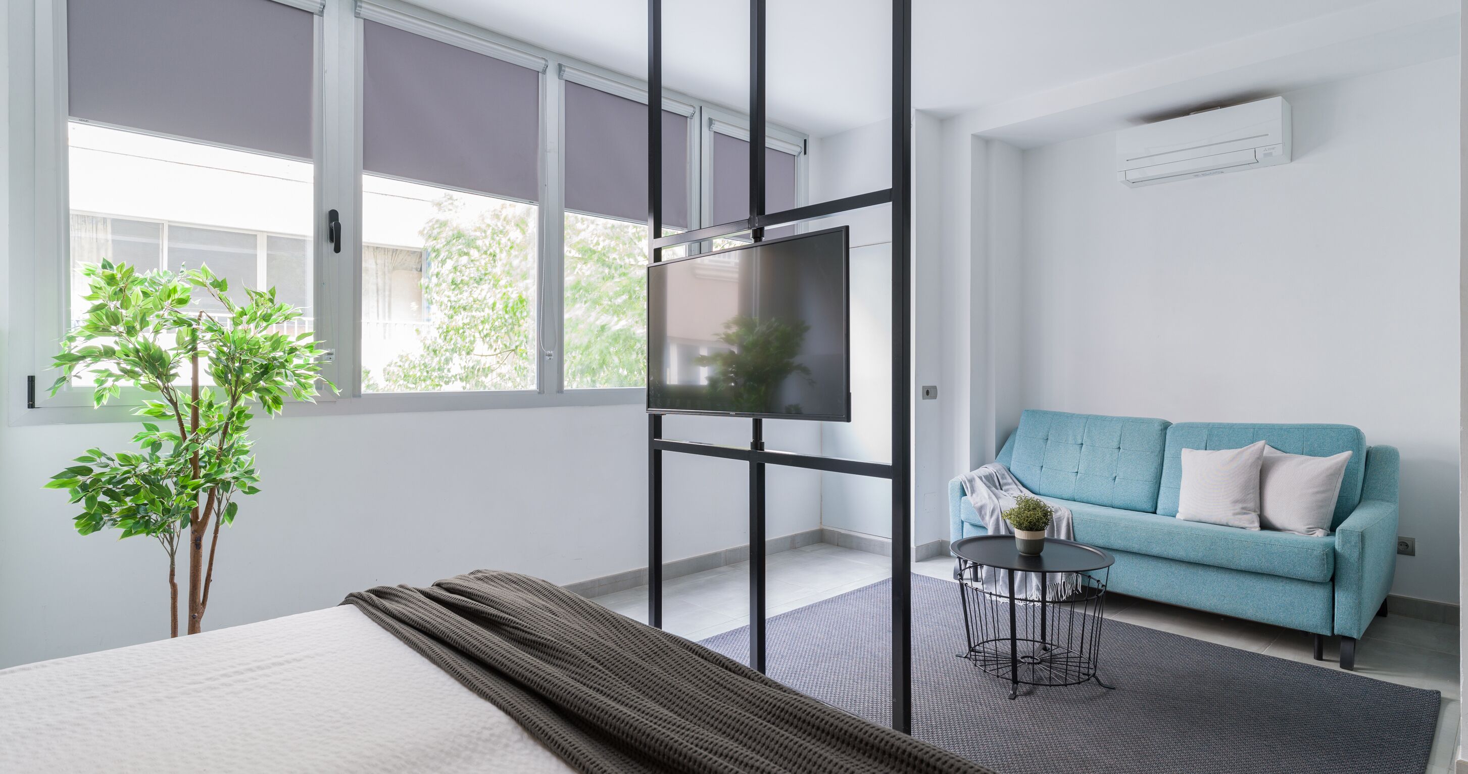 A modern hotel room with a large bed, a small round table, a blue sofa, and a potted plant.