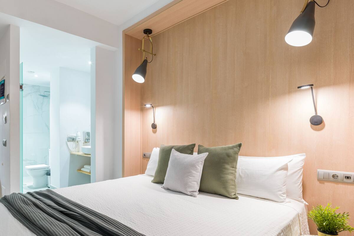 A modern hotel room with a bed, bedside table, and a small potted plant. There is a bathroom visible with a toilet and a sink.