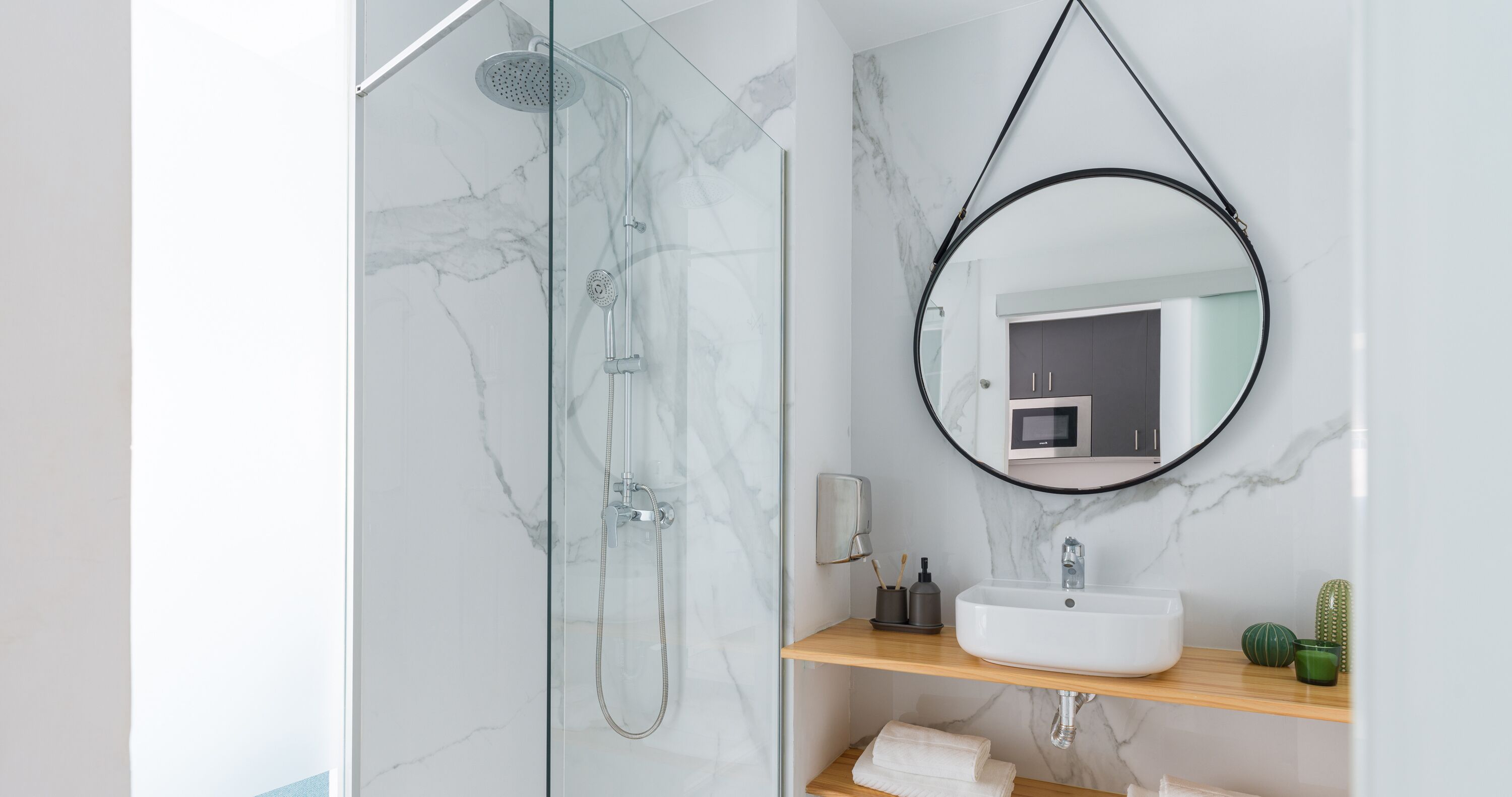 Ein modernes Badezimmer mit einer gläsernen Duschkabine, einem runden Spiegel und einem weißen Waschbecken auf einer Holzablage.