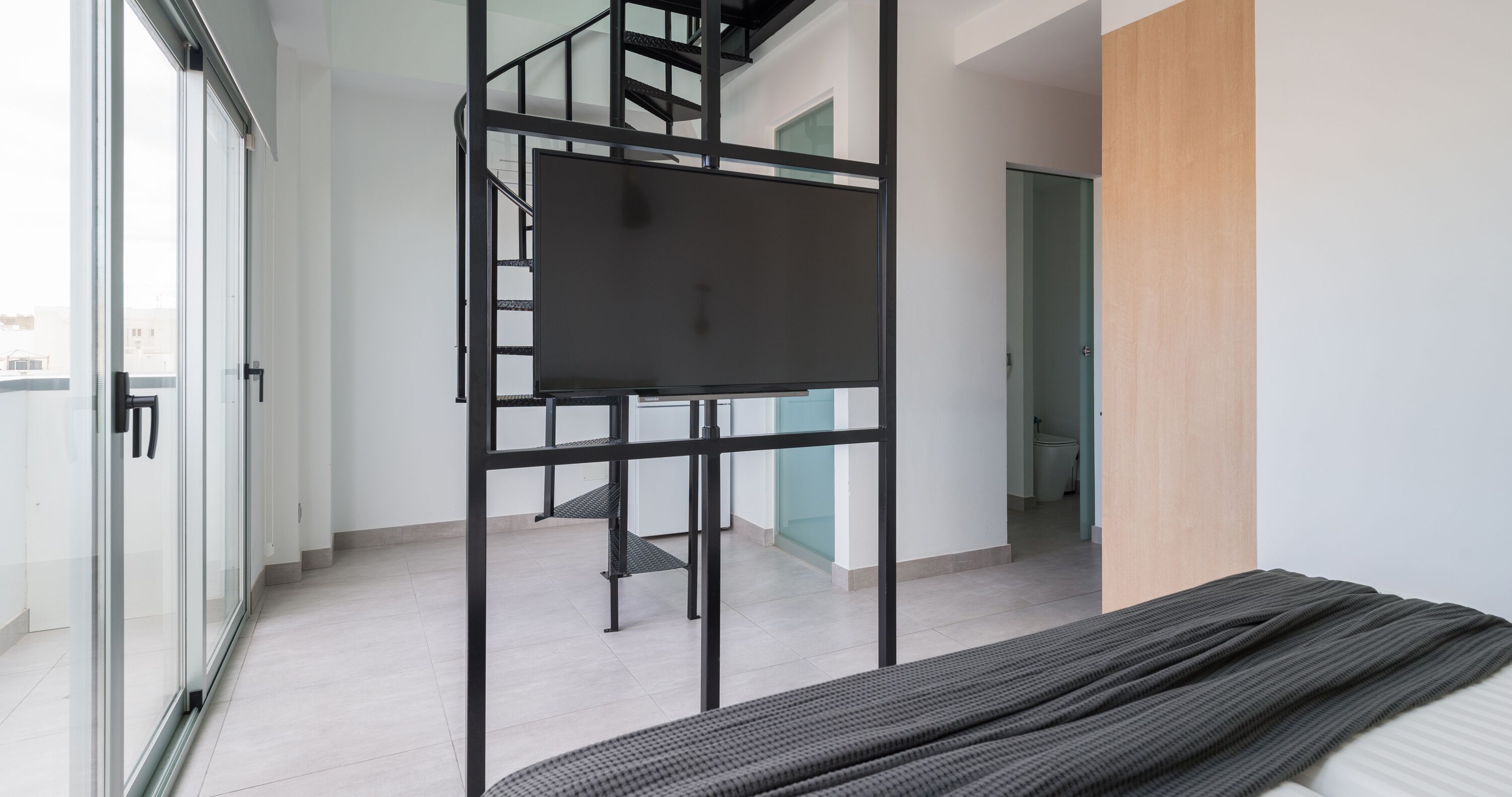 Una habitación moderna con cama, televisor sobre un soporte de metal negro y una escalera que lleva a otra habitación.