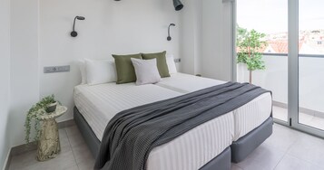 A modern bedroom with a large bed, a small side table with a plant, and a view of the city through a large window.