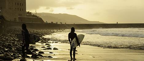 A beach scene with a person carrying a surfboard, a rocky shoreline, and a building in the background.