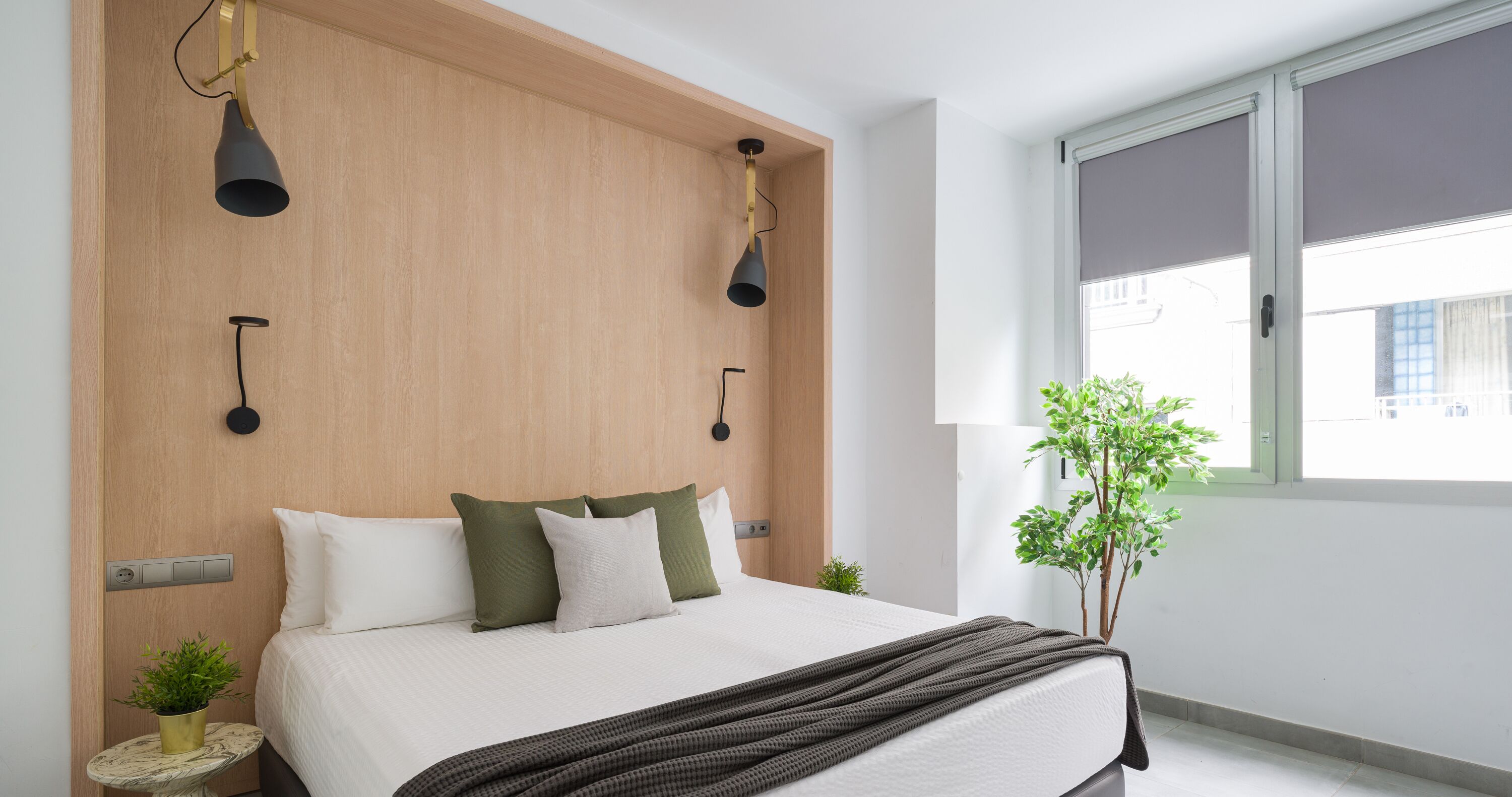 Un dormitorio moderno con cabecera de madera, cama con almohadas blancas y verdes, una manta oscura, una pequeña mesa redonda con una planta y una ventana con vistas a edificios.