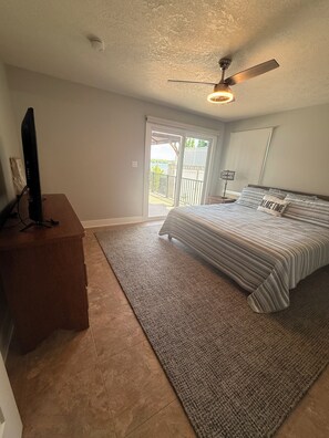 A bedroom with a bed, a TV on a stand, a ceiling fan, and a view through a sliding door to an outdoor area.