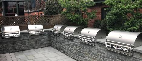 A barbecue area with multiple grills against a stone wall, set in a courtyard with a brick building in the background.