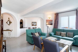 A modern living room with a teal sofa set, a dining table with a patterned tablecloth, and a kitchen area visible through an archway.