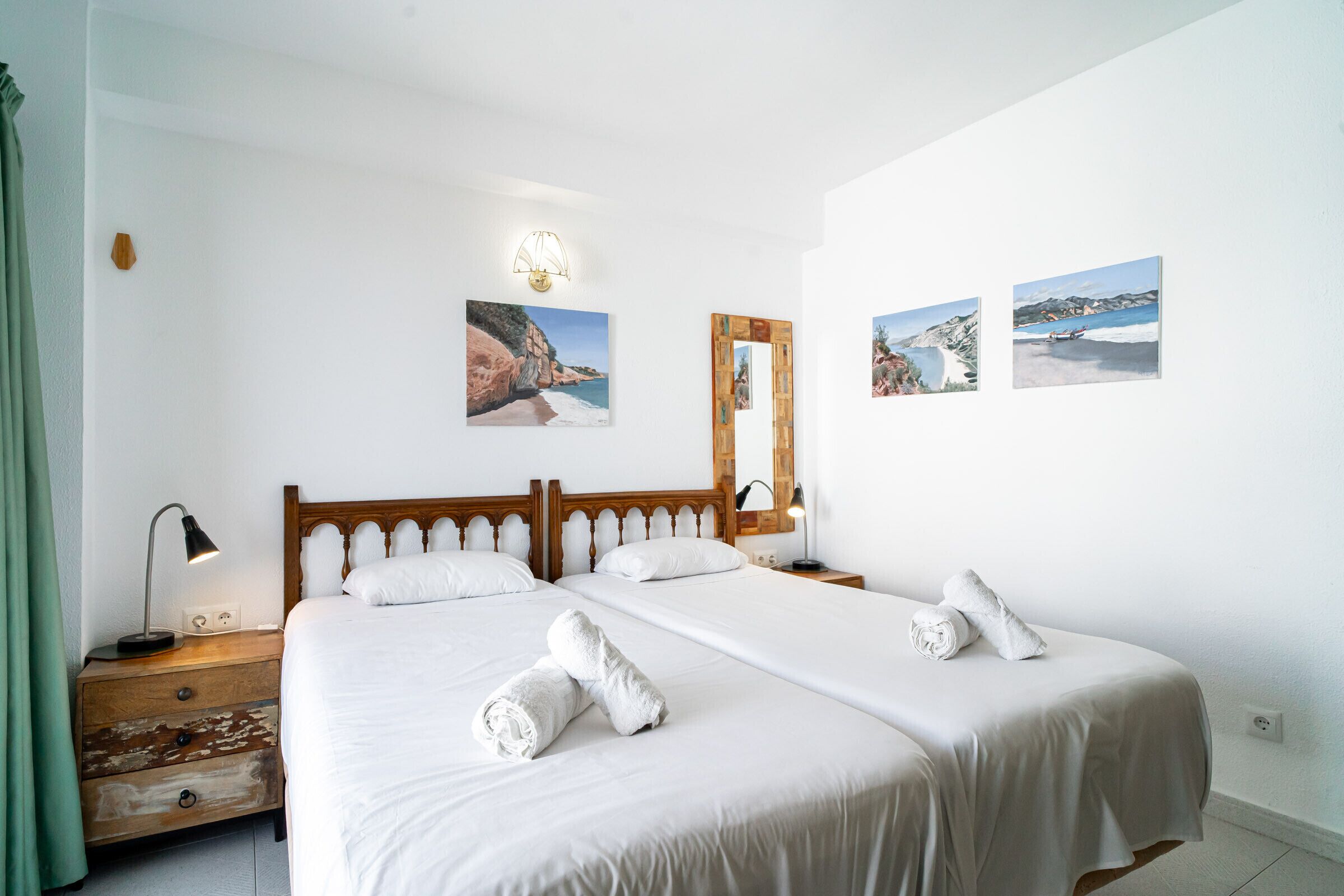 A hotel room with two beds, a wooden headboard, a nightstand with a lamp, a mirror, and artwork on the walls.