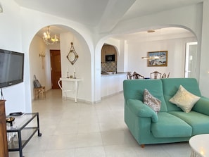 Una sala de estar con un sofá color turquesa, un televisor y un comedor, con una cocina al fondo.