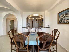 Uma sala de jantar com mesa redonda, seis cadeiras de madeira, toalha de mesa azul e uma luminária pendente moderna.