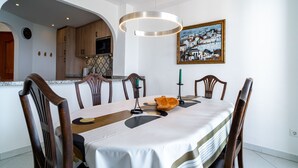 Een eetruimte met een tafel voor vier personen, houten stoelen, een schilderij aan de muur en een keuken zichtbaar door een boog.