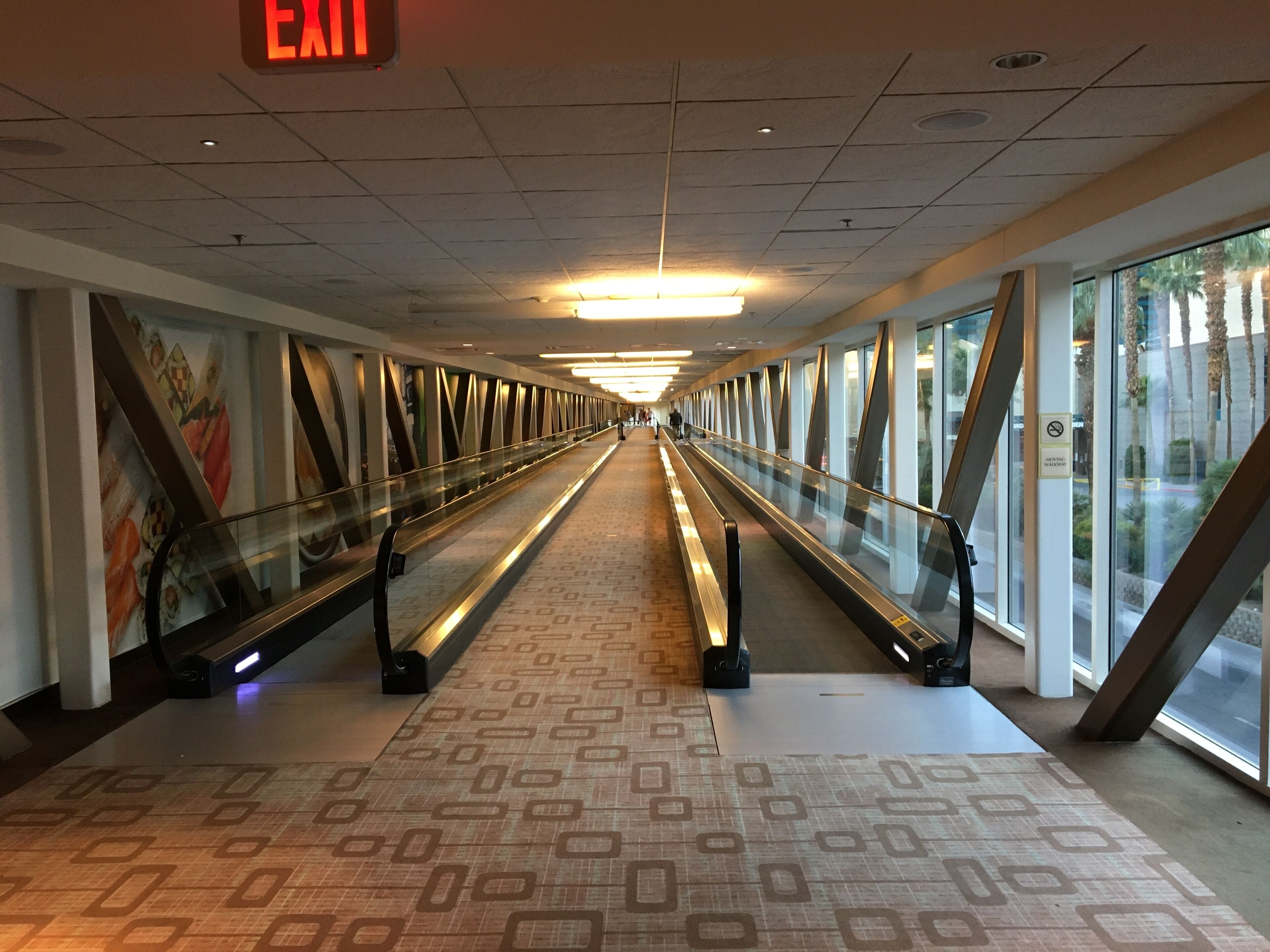 A corridor with escalators, patterned floor, glass railings, and an exit sign.