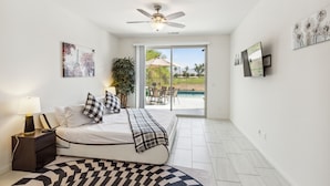 A bedroom with a bed, nightstand, lamp, ceiling fan, and a view of an outdoor patio with a pool and umbrella. - Luxury Golf Retreat w/ Saltwater Pool, Spa &  View, Walk2-Coachella, Stagecoach (Indio)