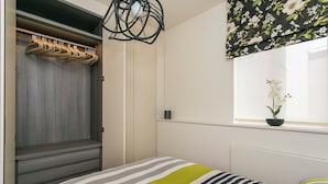 Un dormitorio con cama, armario, lámpara colgante, ventana con planta y radiador.